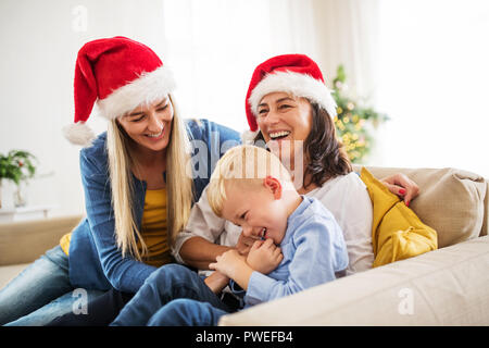 Un piccolo ragazzo con madre e nonna con Santa hat seduto su un divano di casa al tempo di Natale, divertendosi. Foto Stock