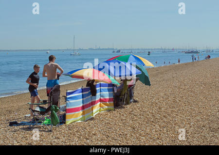 Cowes. L'Isola di Wight. Spiaggia ghiaiosa. Litorale. Adulti sotto ombrello coloratissimo ombrelloni e frangivento. Due ragazzi nel nuoto costumi. Barche e persone. La ricreazione. Vacanze a. ​The Solent. Hampshire. ​England meridionale. Il Regno Unito. Foto Stock