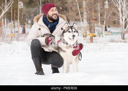 A piena lunghezza ritratto della moderna uomo asiatico che posano con splendidi cani Husky seduti all'aperto nel paesaggio invernale e sorridente felicemente, spazio di copia Foto Stock