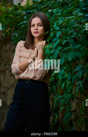 Giovane donna in beige camicetta è in posa lungo una recinzione di pietra coperto con foglie verdi. Foto Stock