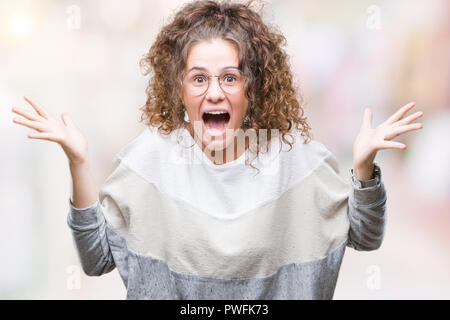 Bella bruna capelli ricci giovane ragazza con gli occhiali su sfondo isolato celebrando crazy e sorpreso per il successo con le braccia sollevate e aprire e Foto Stock