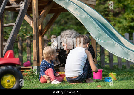 Nonno seduto in un'erba a giocare con i suoi due nipoti in un parco giochi. Foto Stock