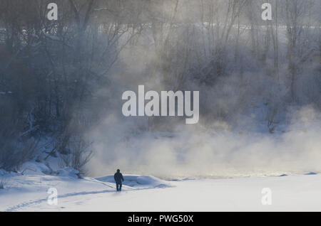 Silhouette di un uomo sulla costa del fiume congelato con la nebbia illuminata dal sole di mattina in inverno Foto Stock