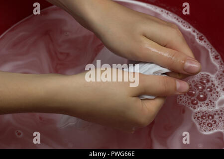 La ragazza cancella un bambini T-shirt bianco in un bacino di rosso da macchie, pulisce da uno smacchiatore, close-up, mani, sfondo bianco, pulizia Foto Stock