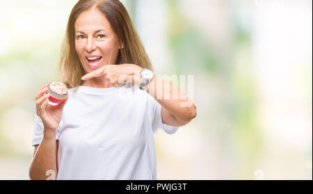 Medioevo donna ispanica mangiare cupcake su sfondo isolato molto felice puntando con mani e dita Foto Stock