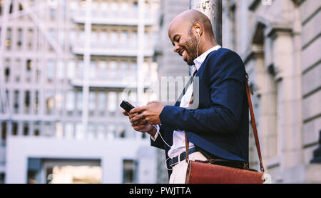 Felice giovane imprenditore con uno smartphone all'aperto a piedi in strada della citta'. L'uomo africano in tuta con gli auricolari per ascoltare musica su un telefono cellulare fuori Foto Stock