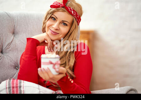 Giovani sorridente bella donna in poltrona con una tazza di cioccolato caldo avvolto in una coperta durante il tempo di Natale Foto Stock