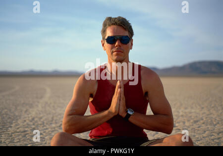 Calma giovane uomo che indossa gli occhiali da sole e la seduta con le gambe incrociate meditando in un deserto remoto. Foto Stock