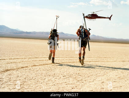 Avventurosa coppia giovane trasportare attrezzature sportive sulle loro spalle sotto hovering elicottero mentre si esegue attraverso un deserto. Foto Stock