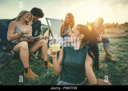 Felice giovani amici di godere di una giornata di sole in montagna. Stanno a ridere e cantare con la musica di ukulele vicino alla tenda. Foto Stock