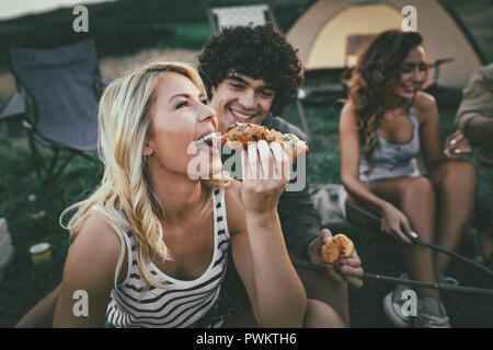 Felice coppia giovane godere di una giornata di sole in compagnia di amici in montagna. Stanno ridendo e mangiando salsicce alla griglia vicino alla tenda. Foto Stock