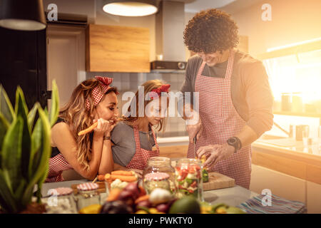 Carino bambina e la sua bella i genitori sono il taglio di vegetali e di sorridere mentre rendendo pickle in cucina a casa Foto Stock