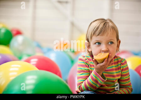Piccola ragazza seduta in una stanza piena di palloncini e mangiare una tortina. Foto Stock