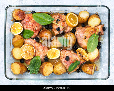 Vista dall'alto di cosce di pollo con patate, limone e olive nere, cotti in forno su cemento grigio Sfondo. Il pollo al forno gamba trimestre in calore-prova di vetro. Foto Stock