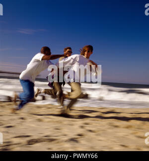 Tre giovani uomini la riproduzione di football americano insieme su una spiaggia di sabbia al sole. Foto Stock