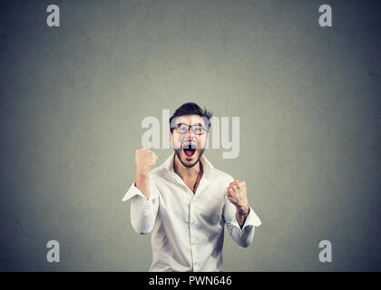 Super eccitati giovane uomo in bicchieri tenendo i pugni nella celebrazione di successo su sfondo grigio Foto Stock