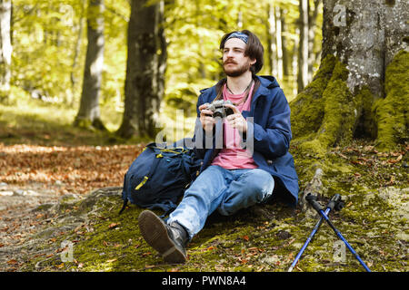 Il lato orizzontale vista ritratto di giovani caucasici brunette uomo barbuto che indossa t-shirt rosa, mantello blu jeans e tenere la fotocamera e meditando solo rilassante su MOSS front di albero circondato da foresta, stile di vita viaggio concetto di successo Avventura Estate attiva vacanze outdoor sport alpinismo Foto Stock