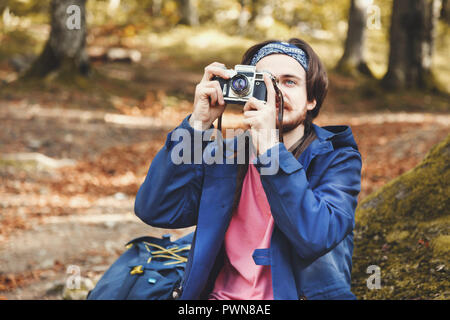 Il lato orizzontale vista ritratto di bello, barbuto ragazzo indossa t-shirt rosa, mantello blu e jeans con lo zaino e tenere la fotocamera e scattare le foto di autunno foresta su MOSS front di albero circondato da foreste, camp, avventura, viaggio, concetto Foto Stock