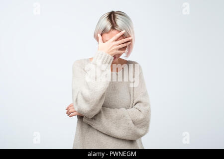 Giovane donna europea in grigio maglione nasconde il suo volto, studio foto isolato su sfondo grigio. Ella ha fobia sociale, cercando di essere anonym Foto Stock