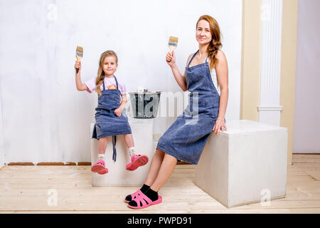 Riparazione nell'appartamento. La famiglia felice la madre e la figlia in grembiuli preparato per dipingere la parete con vernice bianca. Sedersi con fiocchi nelle loro mani Foto Stock