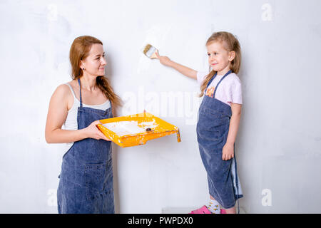 Riparazione nell'appartamento. La famiglia felice la madre e la figlia in grembiuli dipingere la parete con vernice bianca. la figlia dipinge la parete con un pennello e la madre mantiene la cella di vernice e osserva Foto Stock