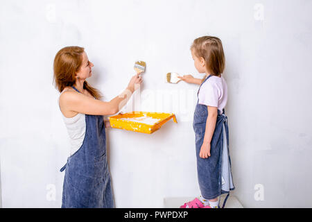 Riparazione nell'appartamento. La famiglia felice la madre e la figlia in grembiuli dipingere la parete con vernice bianca. la figlia dipinge la parete con un pennello e la madre mantiene la cella di vernice e aiuta a Foto Stock