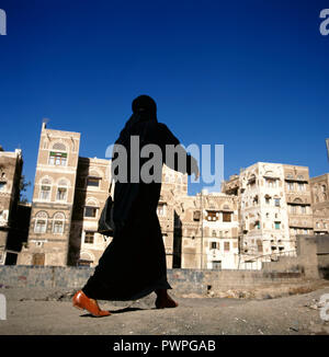 Una donna musulmana velata passeggiate su un Sanaa street, Yemen.a sfondo tipiche case dello Yemen. Foto Stock