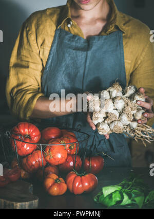 Donna in lino grembiule holding mazzetto di aglio per la cottura di salsa di pomodoro, pomodori pelati o pasta con basilico e pomodori cimelio di famiglia al banco di cucina. H Foto Stock