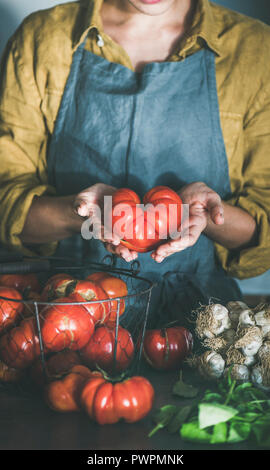 Donna in lino grembiule tenendo mature cimelio di pomodoro per la cottura di salsa di pomodoro, pomodori pelati o pasta con basilico e aglio in cucina. Un sano Foto Stock