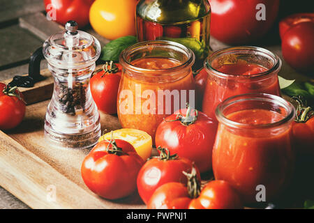 Varietà di tre fatti in casa salse di pomodoro in vasi di vetro con gli ingredienti di cui sopra. Diversi tipi di pomodoro, basilico, olio d'oliva, pepe, sale nel vassoio di legno Foto Stock