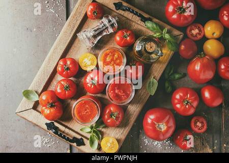 Varietà di tre fatti in casa salse di pomodoro in vasi di vetro con gli ingredienti di cui sopra. Diversi tipi di pomodoro, basilico, olio d'oliva, pepe, sale nel vassoio di legno Foto Stock