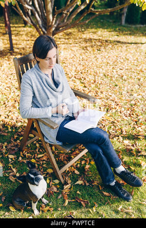 Donna che indossa caldo abbigliamento in maglia di bere una tazza di tè o caffè caldo e la lettura di un libro all'aperto alla luce del sole - Stagione, letteratura, tempo libero e pe Foto Stock