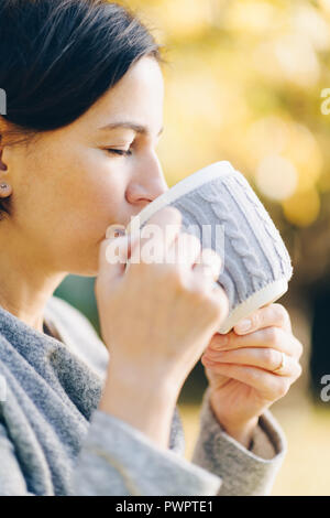 Donna che indossa caldo abbigliamento in maglia di bere una tazza di tè o caffè caldo all'aperto alla luce del sole - stagione, il tempo libero e il concetto di persone - una bella mattina Foto Stock