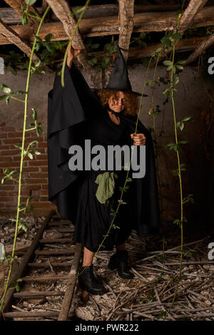 Vecchio scary strega in una casa in rovina, vestito di nero Foto Stock