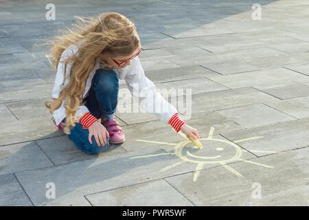 Bambino ragazza richiama il simbolo del sole con le matite colorate su asfalto. Foto Stock