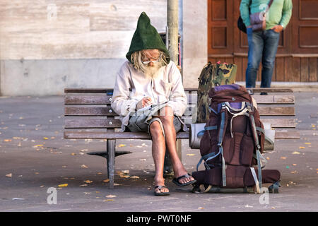 Ancona, Italia - Settembre 27th, 2018: un uomo anziano backpacker seduta su una panchina quadrato, è scrivere note su un blocco note che indossa occhiali e un Foto Stock
