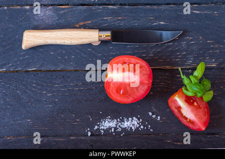 Coltello con un manico in legno e una lama di nero. Fette di pomodori rossi su sfondo nero. Coltello e verdure. Foto Stock