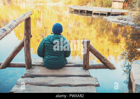 Un uomo seduto su di un molo in legno dal lago in autunno e guarda all'acqua Foto Stock