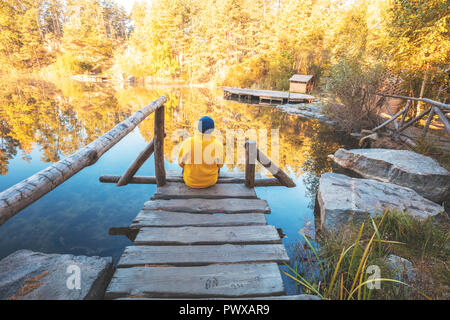 Un uomo seduto su di un molo in legno dal lago in autunno e guarda all'acqua Foto Stock