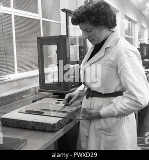 Degli anni Cinquanta, storico, fabbricazione di vernice, un tecnico femmina indossando un white-coat in corrispondenza di un banco di lavoro tenendo un vetro dello schermo e lama di miscelazione, Inghilterra, Regno Unito. Foto Stock