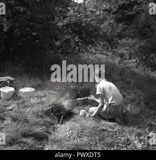 Degli anni Cinquanta, storico, camping, giovane ragazzo cucinando fuori nell'aperto su un prato area piana in una zona appartata riparata una parte di un bosco o di una foresta, Inghilterra, Regno Unito. Foto Stock