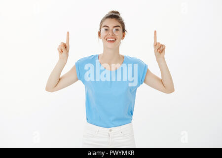 Ritratto di positivo europeo affascinante studentessa di bun acconciatura e bicchieri, sorridente con gioia mentre sollevando le dita indice e rivolta verso l'alto, essendo lieti e soddisfatti con sorprendente rainbow Foto Stock