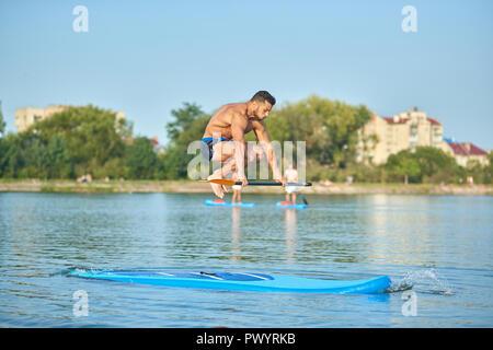 Bel uomo muscolare formazione sulla scheda paddle, saltando su di esso con lungo remo in mano al lago della città. Montare giovane ragazzo indossa pantaloncini sport, esercitando sull'acqua. Concetto di sport e stile di vita sano. Foto Stock