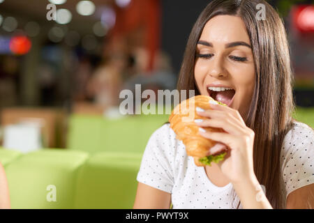 Fame client femmina in seduta cafe e mordere di deliziosi croissant con verdi e prosciutto. Bella bruna in camicia bianca con cena e mangiare il cibo gustoso. Concetto di golosa merenda. Foto Stock