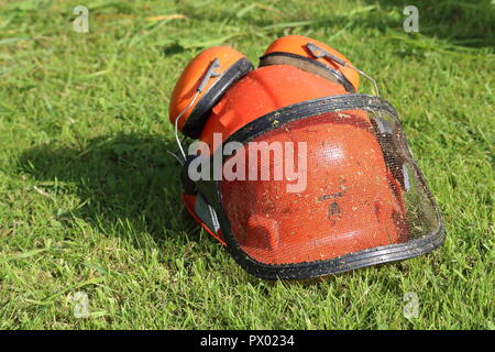 Arancione casco di sicurezza hat con cuffie antirumore, protettori, mesh visiera, erba tagliata dello sfondo. Utilizzato per strimming, giardinaggio, erba sul casco, vista frontale Foto Stock