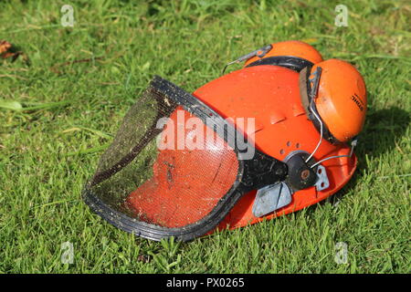 Arancione casco di sicurezza hat con cuffie antirumore, protettori, mesh visiera, erba tagliata dello sfondo. Utilizzato per strimming, giardinaggio, erba sul casco, lato sinistro Foto Stock