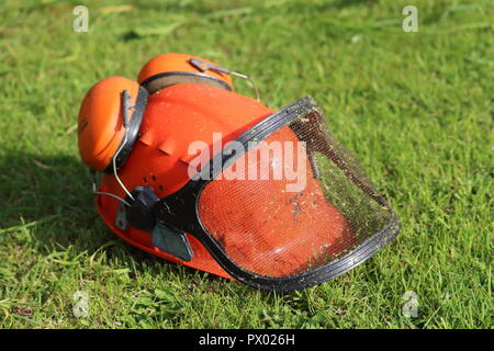 Arancione casco di sicurezza hat con cuffie antirumore, protettori, mesh visiera, erba tagliata dello sfondo. Utilizzato per strimming, giardinaggio, erba sul casco, lato destro Foto Stock