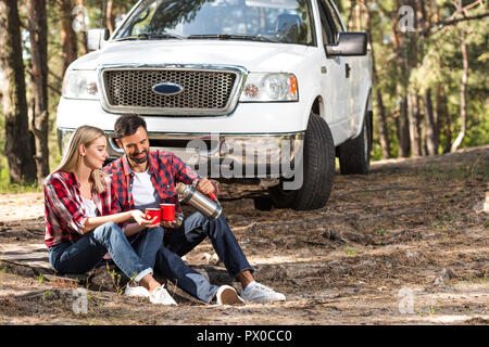 Uomo sorridente versando il caffè alla ragazza tazza da thermos vicino a ritirare l'auto all'aperto Foto Stock