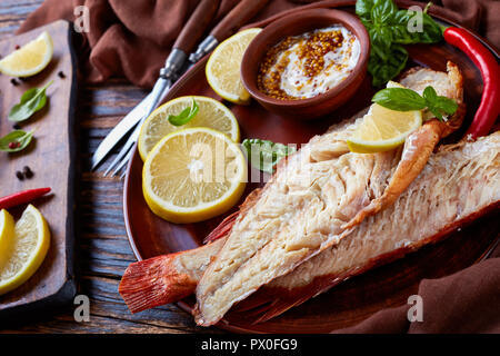 Filetto di red snapper servita su un piatto di terracotta con foglie di basilico, le fette di limone e la salsa di senape, forchetta e coltello su un di legno tavolo rustico, Foto Stock