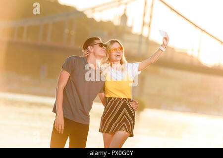 Felice coppia giovane tenendo selfie con lo smartphone sul fiume spiaggia di sera Foto Stock